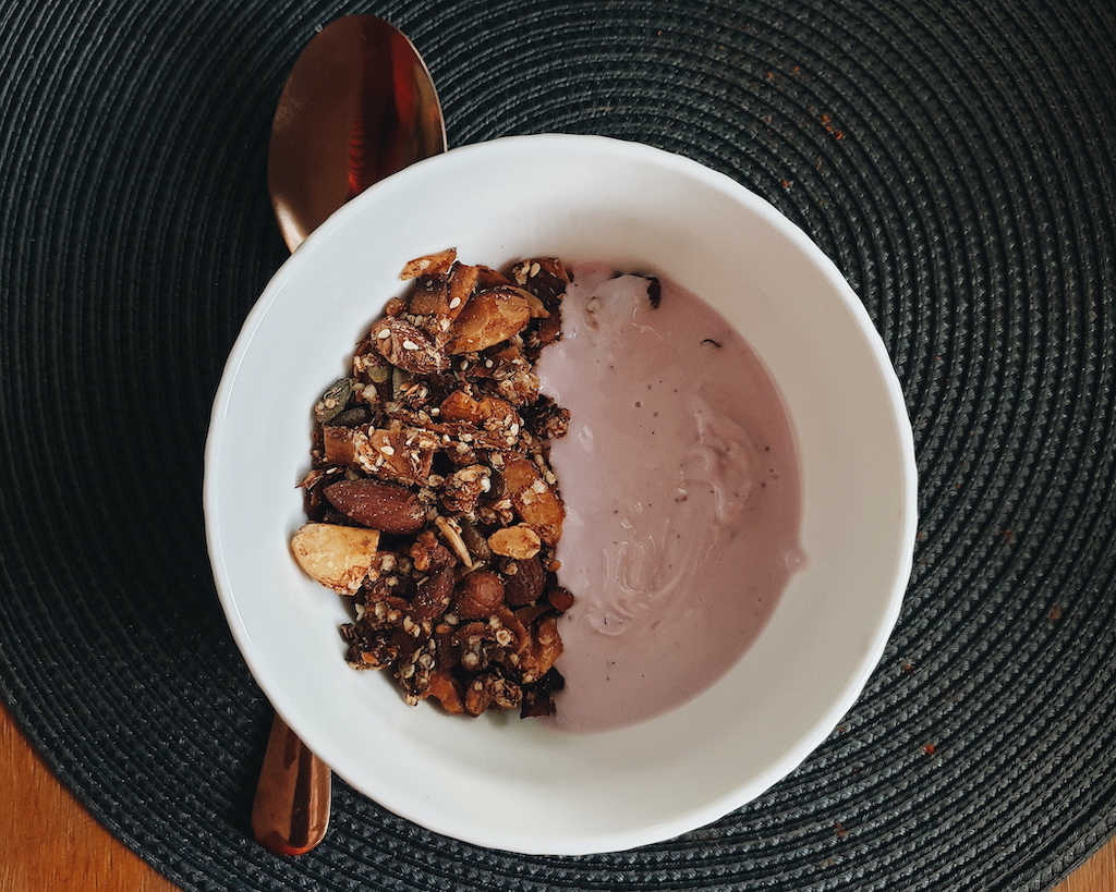 Coconut Grain Free Granola in a white bowl with a bronze spoon. One side of the bowl contains blueberry kefir. 