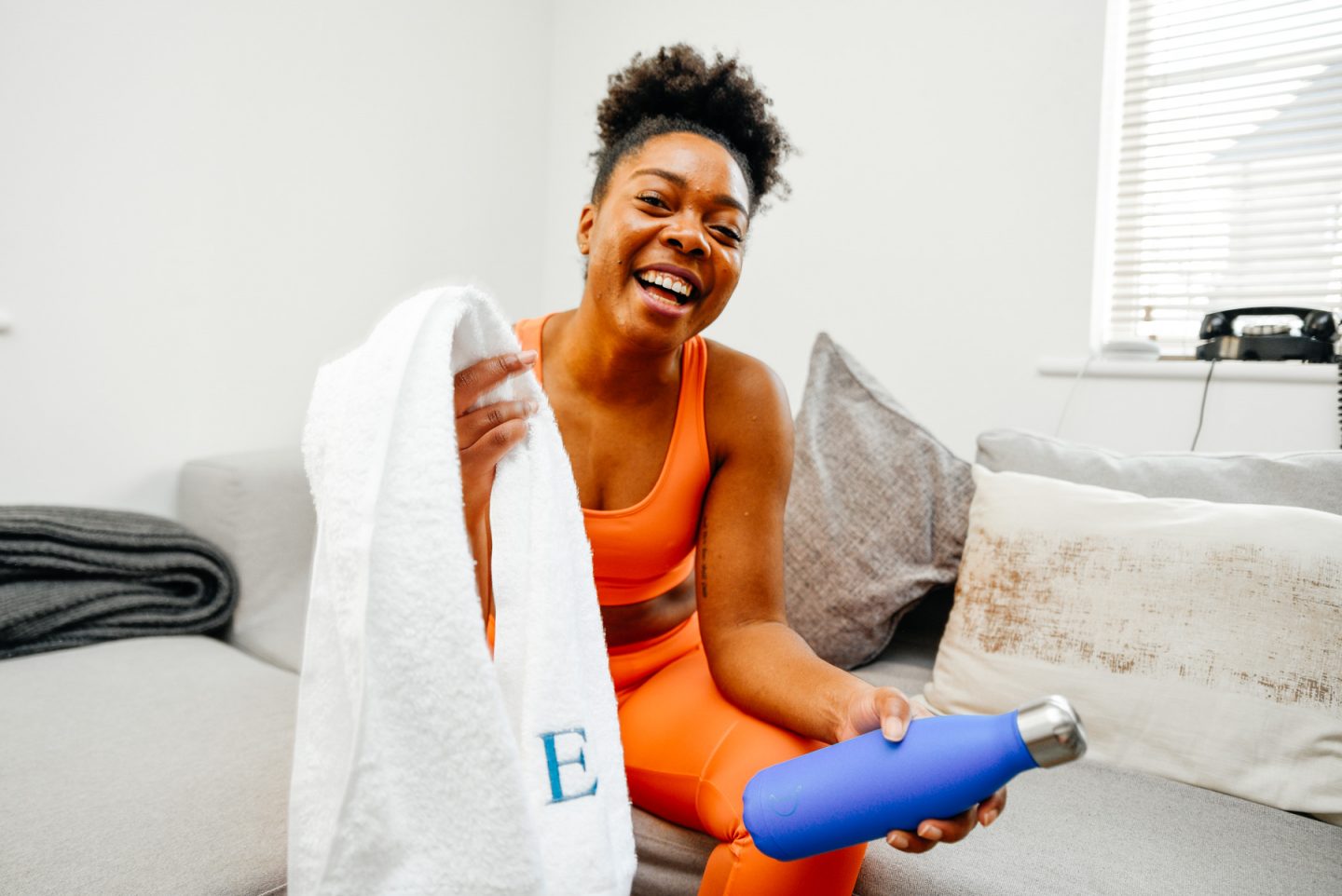 Elle is seated on sofa with white towel and blue water bottle, smiling. Adding Wellbeing Moments To Your Day with Very